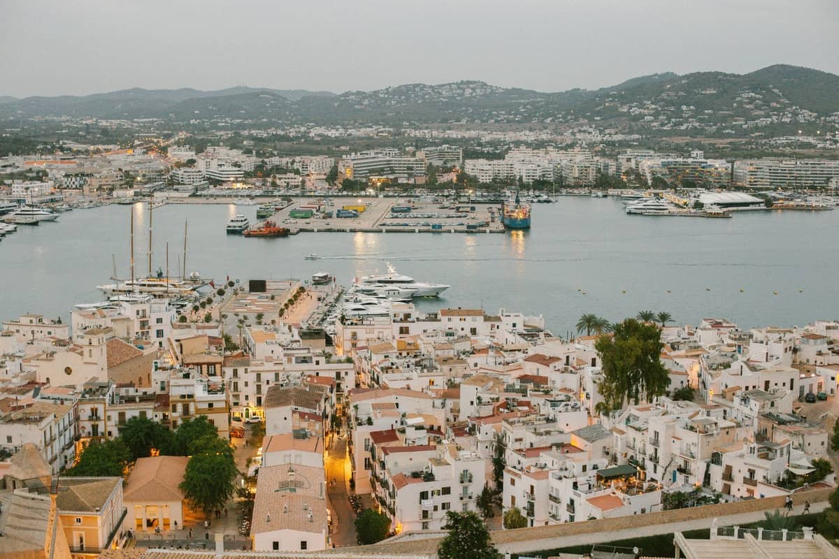 Wide view of Ibiza town and marina with yachts in harbor, white buildings, and hills in the distance at dusk.