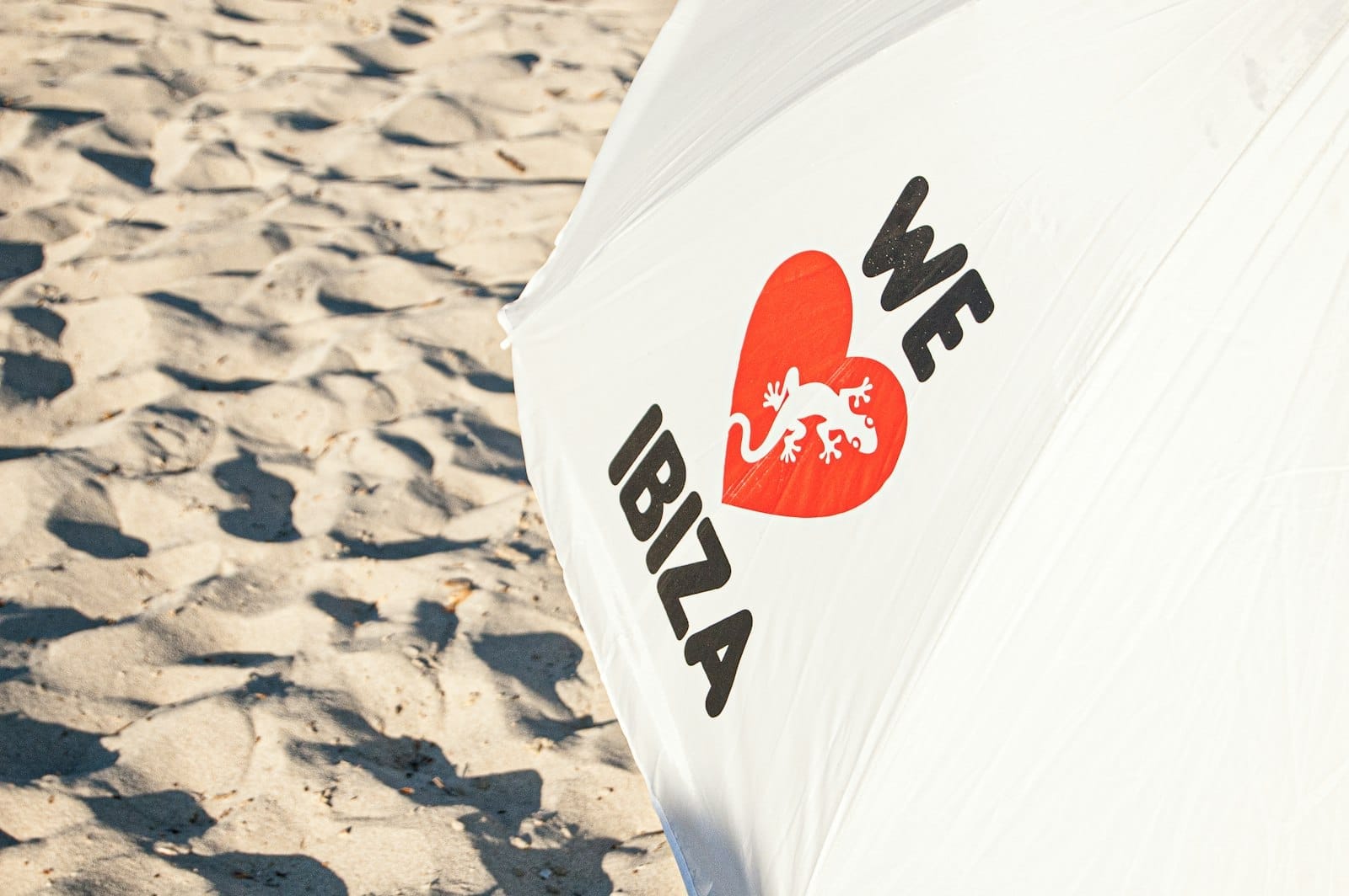 Close-up de um guarda-sol de praia branco com o texto 'We Love Ibiza' e um coração vermelho, sobre fundo de areia de praia.