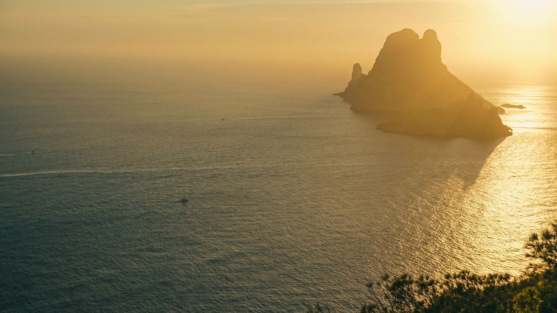 Pôr do sol dourado sobre o mar com a icônica formação rochosa de Es Vedrà em silhueta, vista de um alto penhasco em Ibiza.