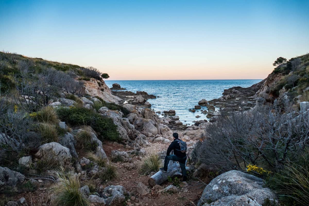 Person with backpack walking along rocky coastal trail towards the sea, with cliffs and sunset sky in Ibiza.