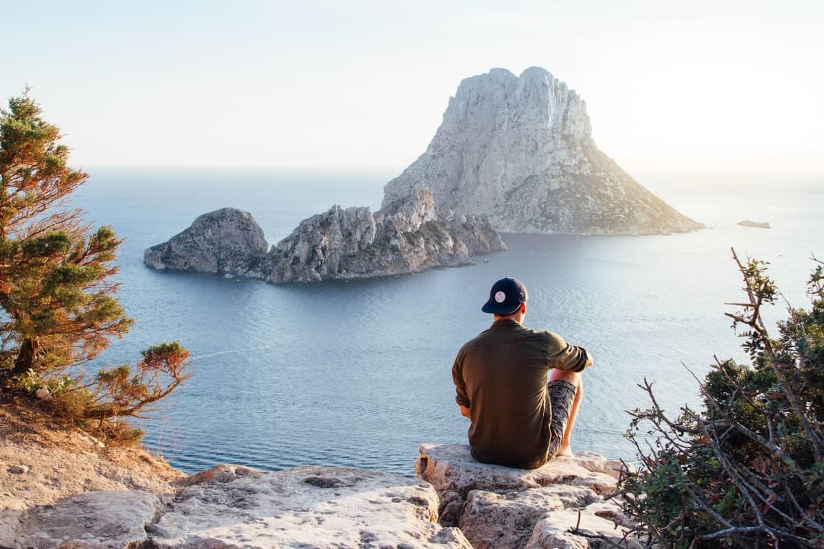 Person sitting on rocky clifftop admiring expansive sea view with iconic Es Vedrà islet under a clear sky.