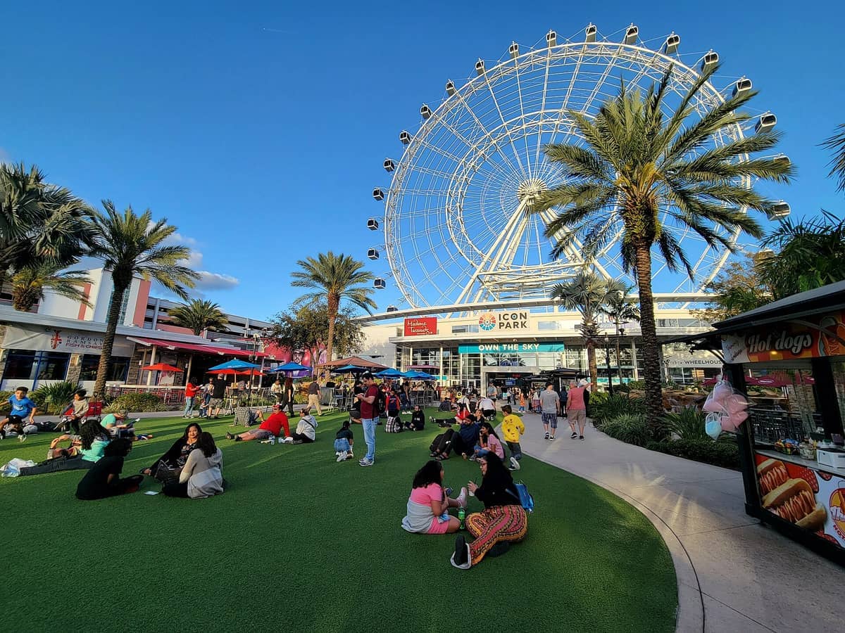 Families and visitors relax on green turf beneath palm trees at ICON Park Orlando, with the towering observation wheel and shops in the background.