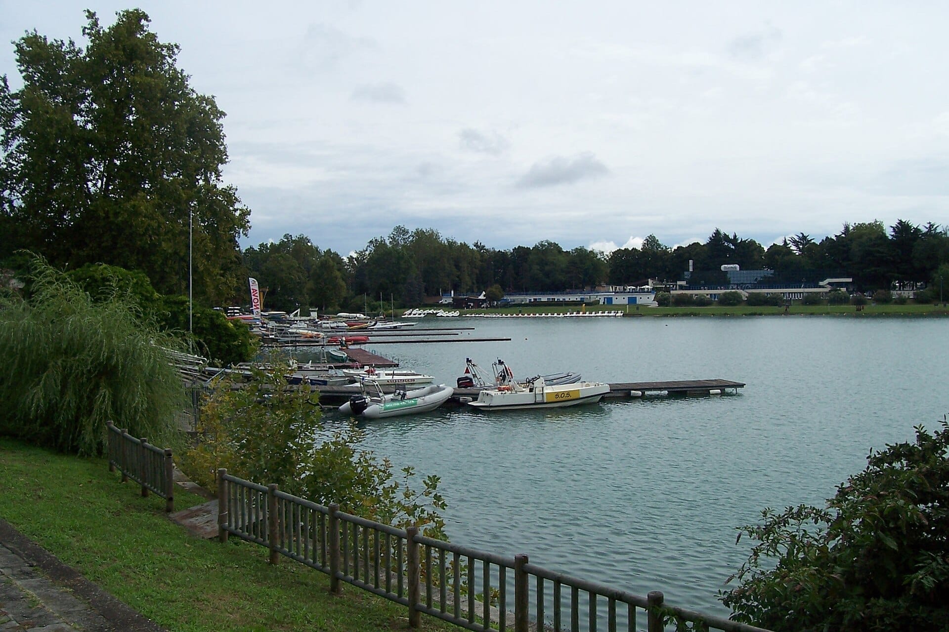 Barche ormeggiate a un molo di legno lungo il lago artificiale alberato dell'Idroscalo di Milano sotto un cielo nuvoloso.