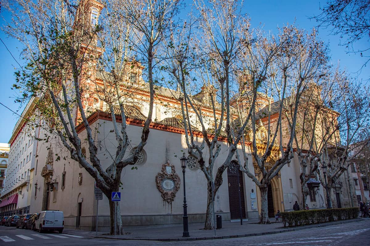 วิวภายนอกของ Iglesia de la Magdalena ในเมือง Seville แสดงให้เห็นส่วนหน้าอาคารสไตล์บาโรกอยู่เบื้องหลังต้นไม้ไร้ใบในวันที่แดดจ้า