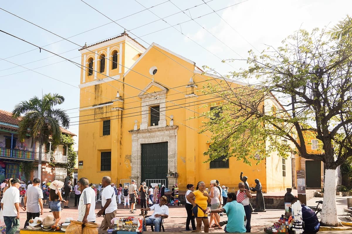 The yellow façade of Iglesia de la Santísima Trinidad stands behind a bustling Plaza de la Trinidad, with people and street vendors under sunny skies.