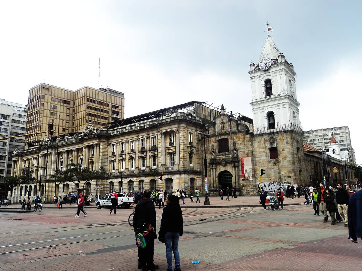 Street-level view of Iglesia de San Francisco in Bogotá, showing its distinctive white clock tower, baroque stone facade, and bustling urban surroundings with pedestrians.
