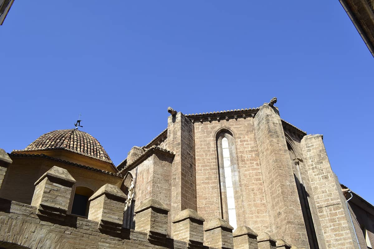 Vista esterna della Iglesia de San Juan del Hospital a Valencia, con le mura in pietra medievale, una cupola rivestita di piastrelle, i contrafforti e il cielo blu sullo sfondo.