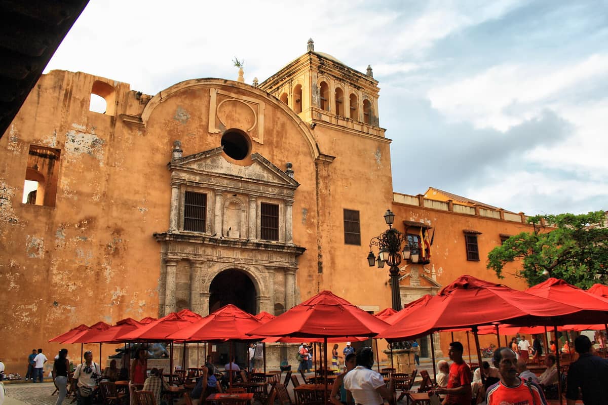 Wide view of Iglesia de Santo Domingo’s historic orange façade and bell tower facing lively plaza with red umbrellas, cafe seating, and people gathered in Cartagena, Colombia.