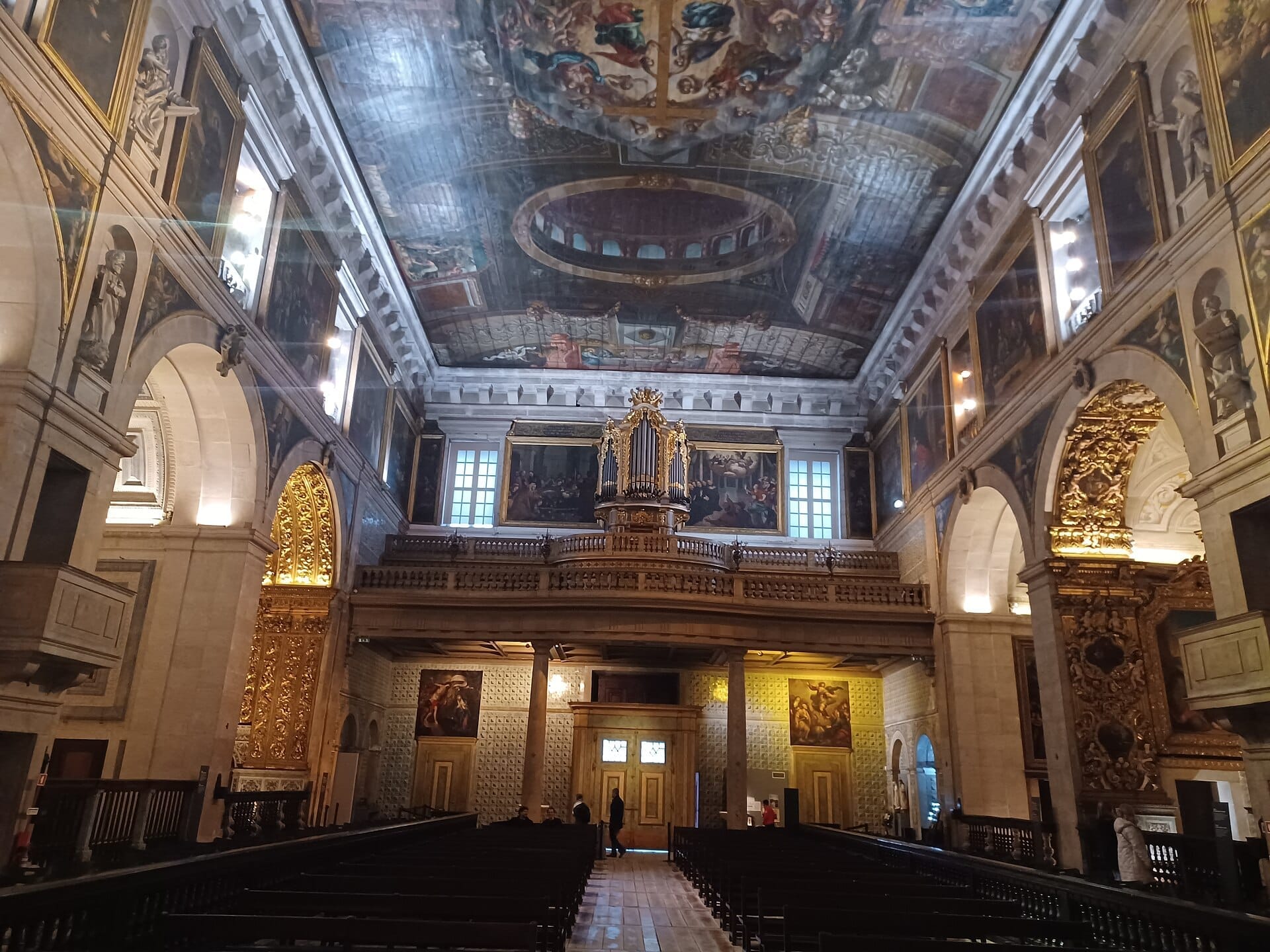 Vista interna della Igreja de São Roque con elaborati affreschi sul soffitto, altari dorati, dipinti classici e banchi in legno in una morbida luce naturale.