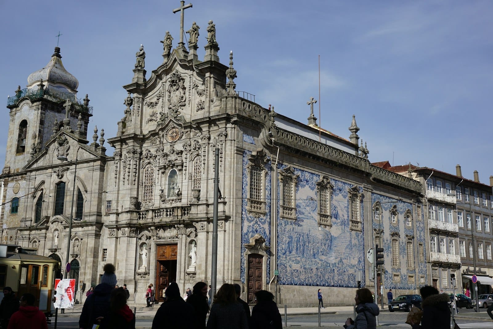 Vista ampia della sontuosa facciata barocca e del famoso pannello laterale di azulejos blu e bianchi dell'Igreja do Carmo, con persone e palazzi intorno in una giornata di sole.