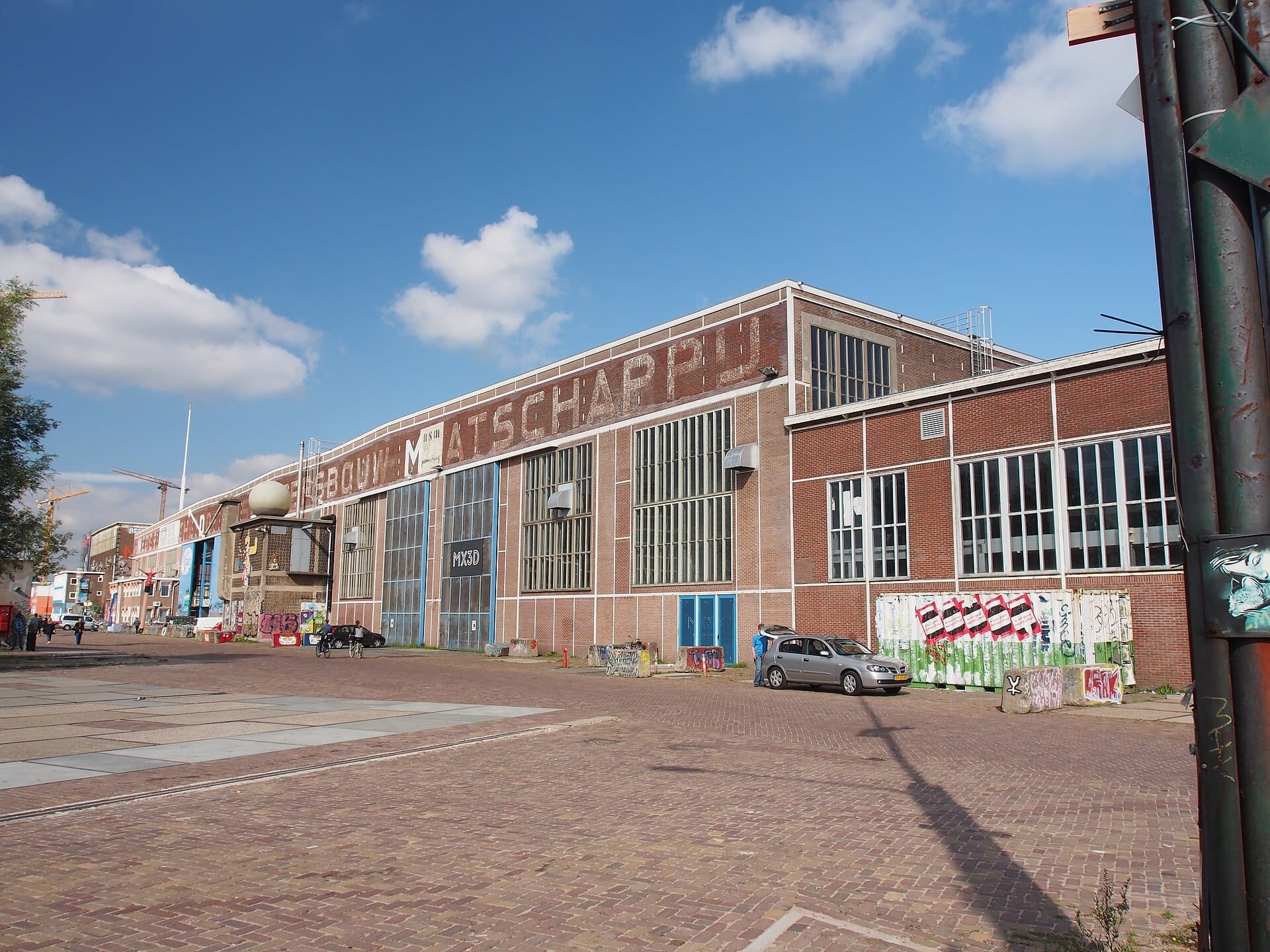 Vista exterior del pabellón industrial NDSM donde se celebra el mercadillo IJ-Hallen, con paredes de ladrillo rojo, grandes ventanas y arte callejero disperso.
