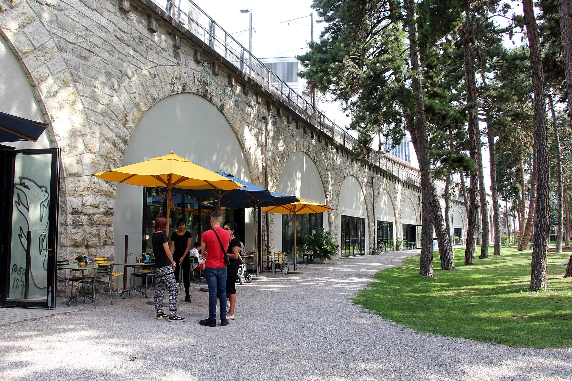 ซุ้มสะพานหินของ Im Viadukt ในซูริค มีร้านกระจกทันสมัยอยู่ใต้ซุ้ม ผู้คนรวมกลุ่มใต้ร่มเหลืองกลางแจ้ง ทางเดินมีต้นไม้เรียงราย
