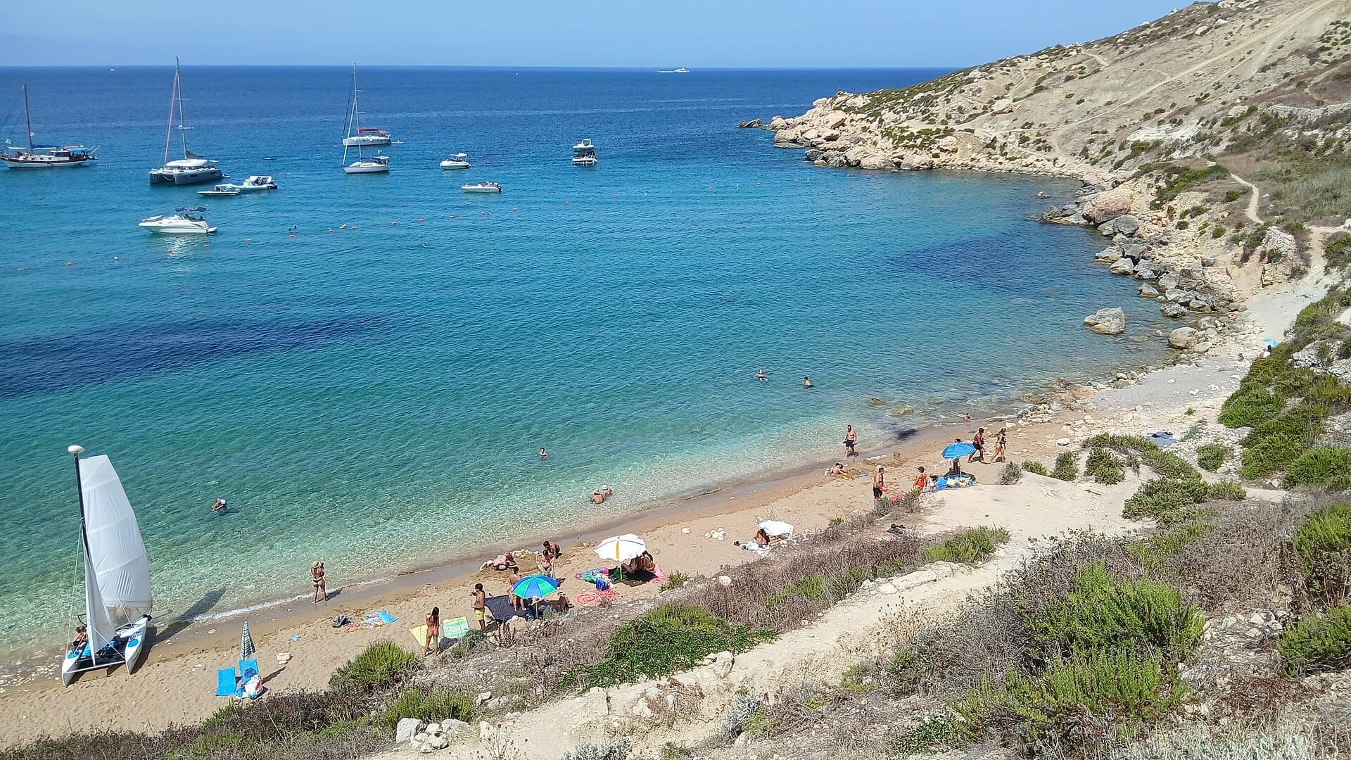 Una distesa di sabbia a Imgiebah Bay con acque turchesi, piccole barche ancorate al largo, bagnanti che prendono il sole, ombrelloni e scogliere rocciose aride sullo sfondo.
