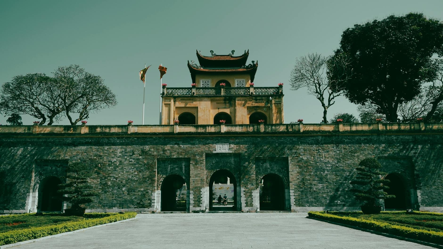 Front view of the main gate and central building of the Imperial Citadel of Thang Long in Hanoi, flanked by flags and trees under a clear sky.