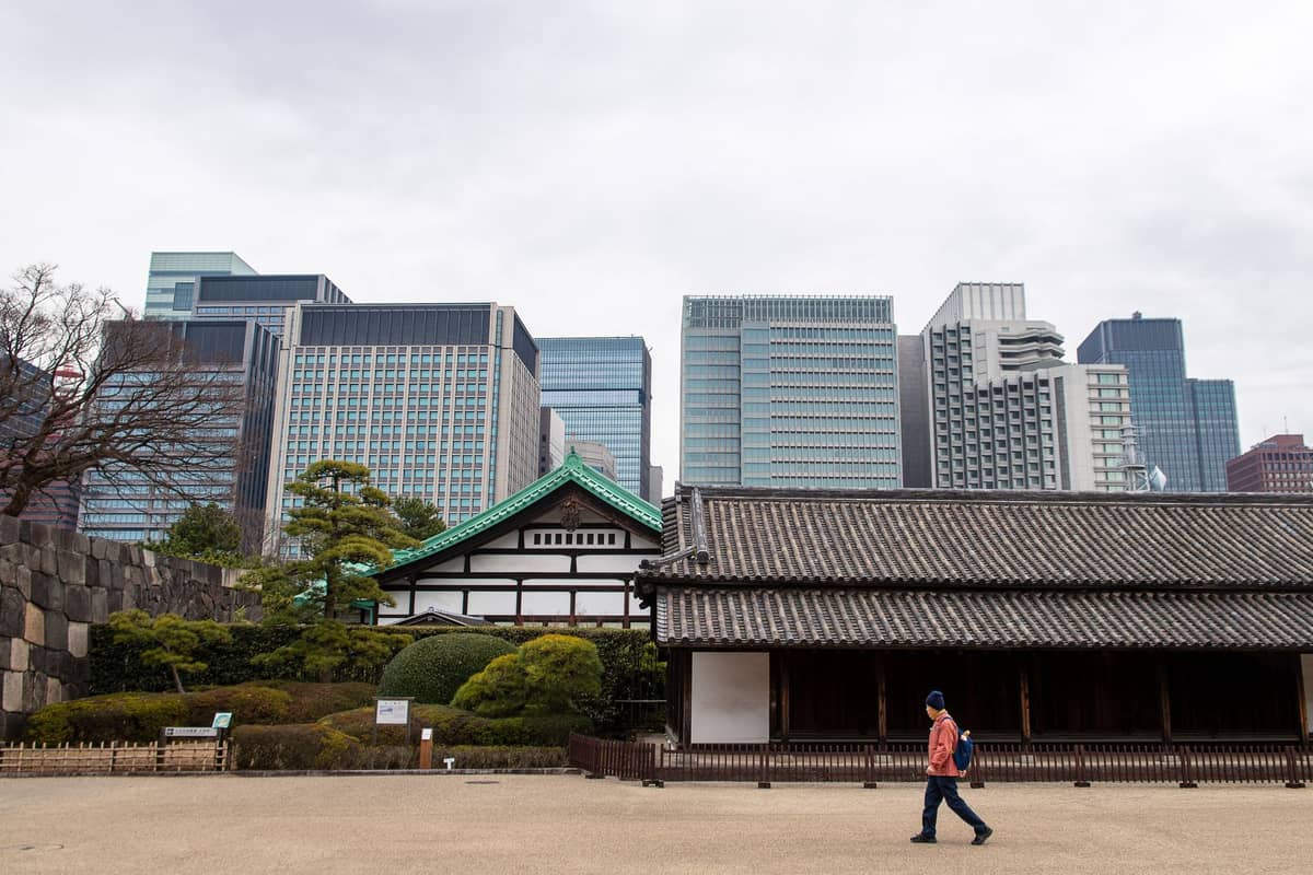 Restored Edo-period buildings and stone walls of the Imperial Palace East Gardens with Tokyo skyscrapers in the background on a cloudy day.
