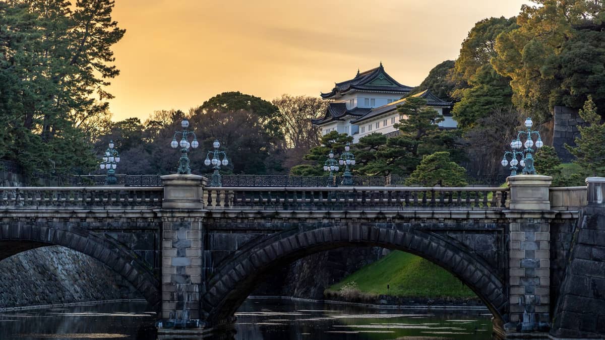 Stone bridge and main gate of Tokyo Imperial Palace with the Fushimi Yagura watchtower in the background at sunset, reflected in the water of the surrounding moat.
