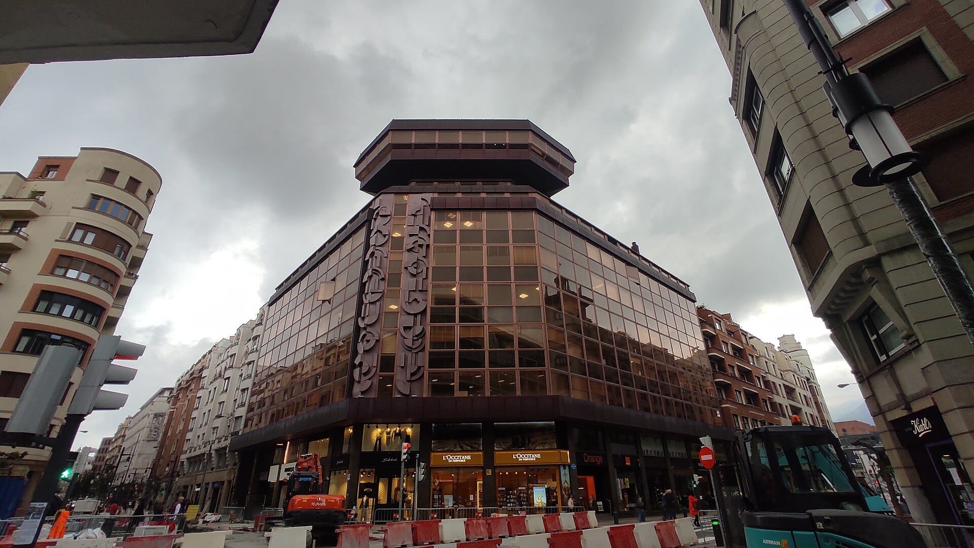 Modern glass building with geometric architecture in Indautxu, Bilbao, under a cloudy sky on a city street lined with shops.