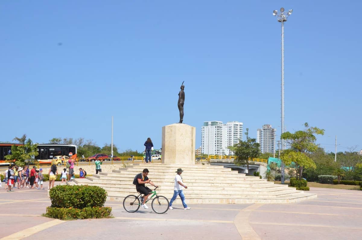 India Catalina Statue on a stone pedestal, surrounded by people, green bushes, and Cartagena’s modern city buildings under a clear blue sky.