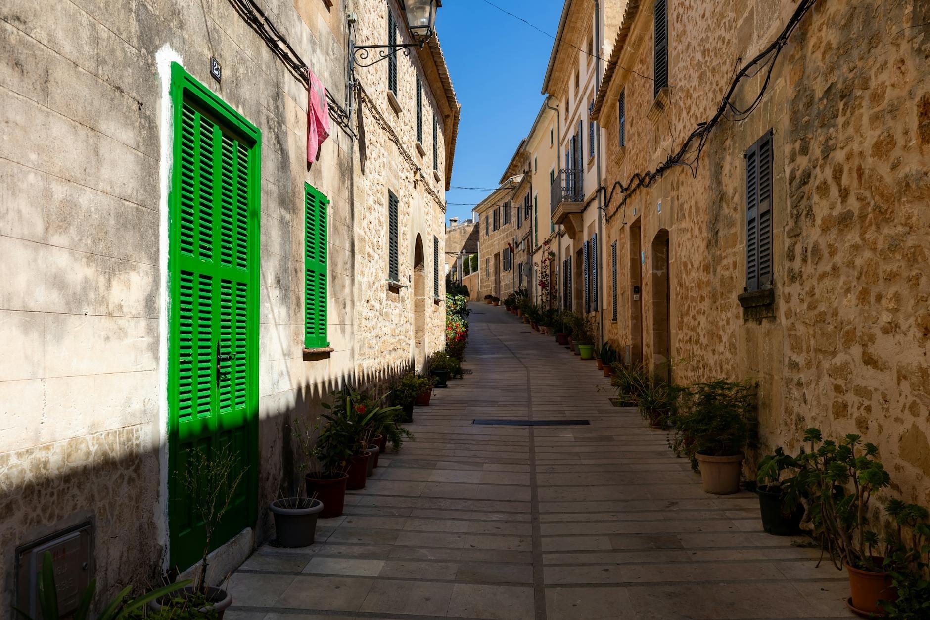 ถนนหินแคบในเขต Es Pla เมือง Mallorca เรียงรายด้วยต้นไม้กระถางและบ้านดั้งเดิมที่มีบานเกล็ดสีเขียวและสีน้ำเงินภายใต้แสงแดดจ้า