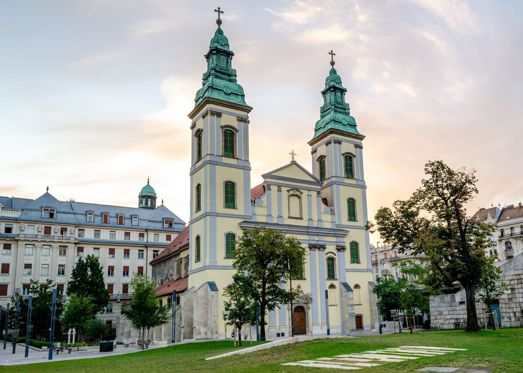 Frontansicht der Innerstädtischen Pfarrkirche in Budapest mit ihren barocken Zwillingstürmen, grünen Dächern und dem umliegenden Platz im Abendlicht.