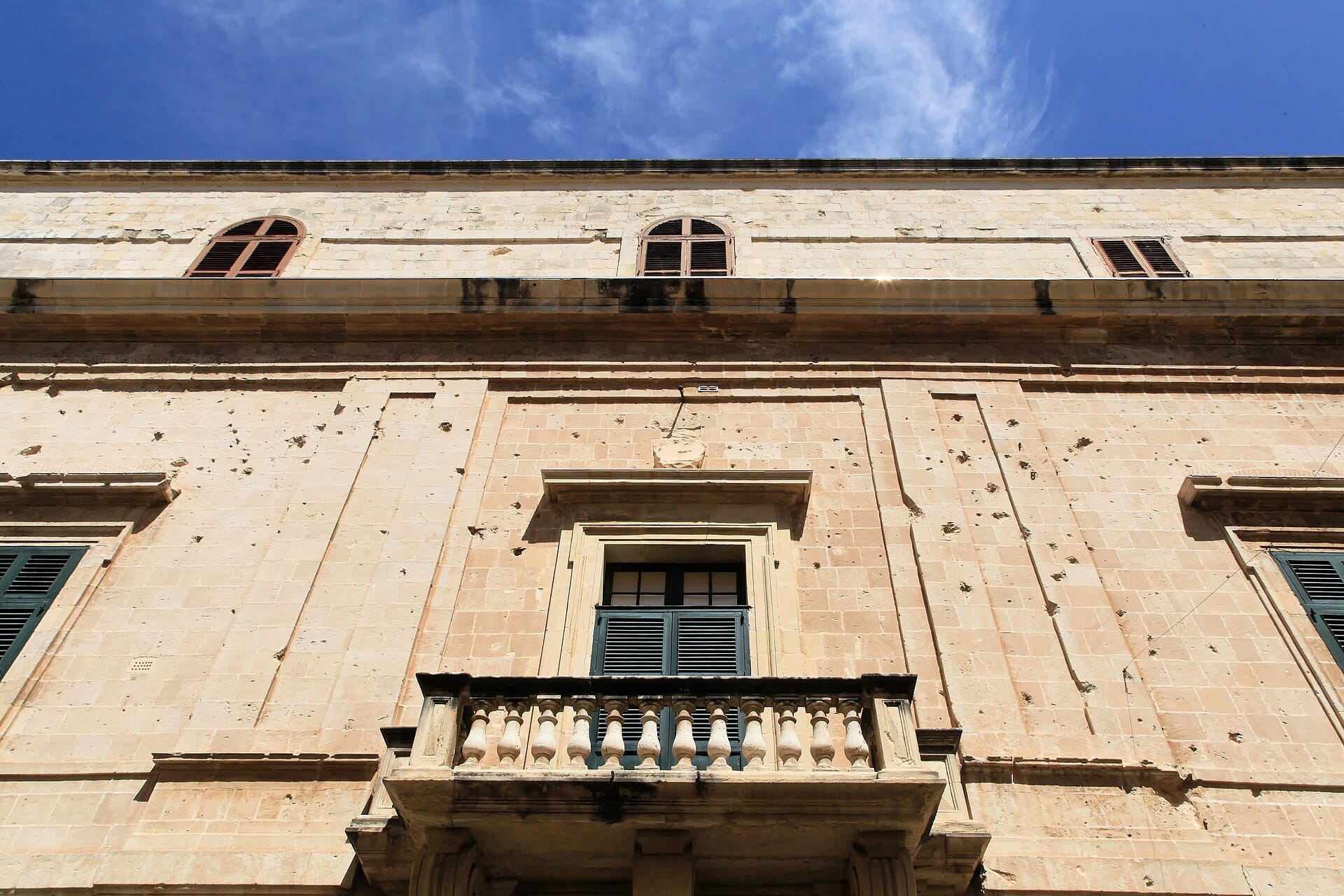 Facciata del Palazzo dell'Inquisitore a Birgu, con mura in pietra calcarea consumata, finestre con persiane e un balcone in pietra sotto un cielo azzurro limpido.