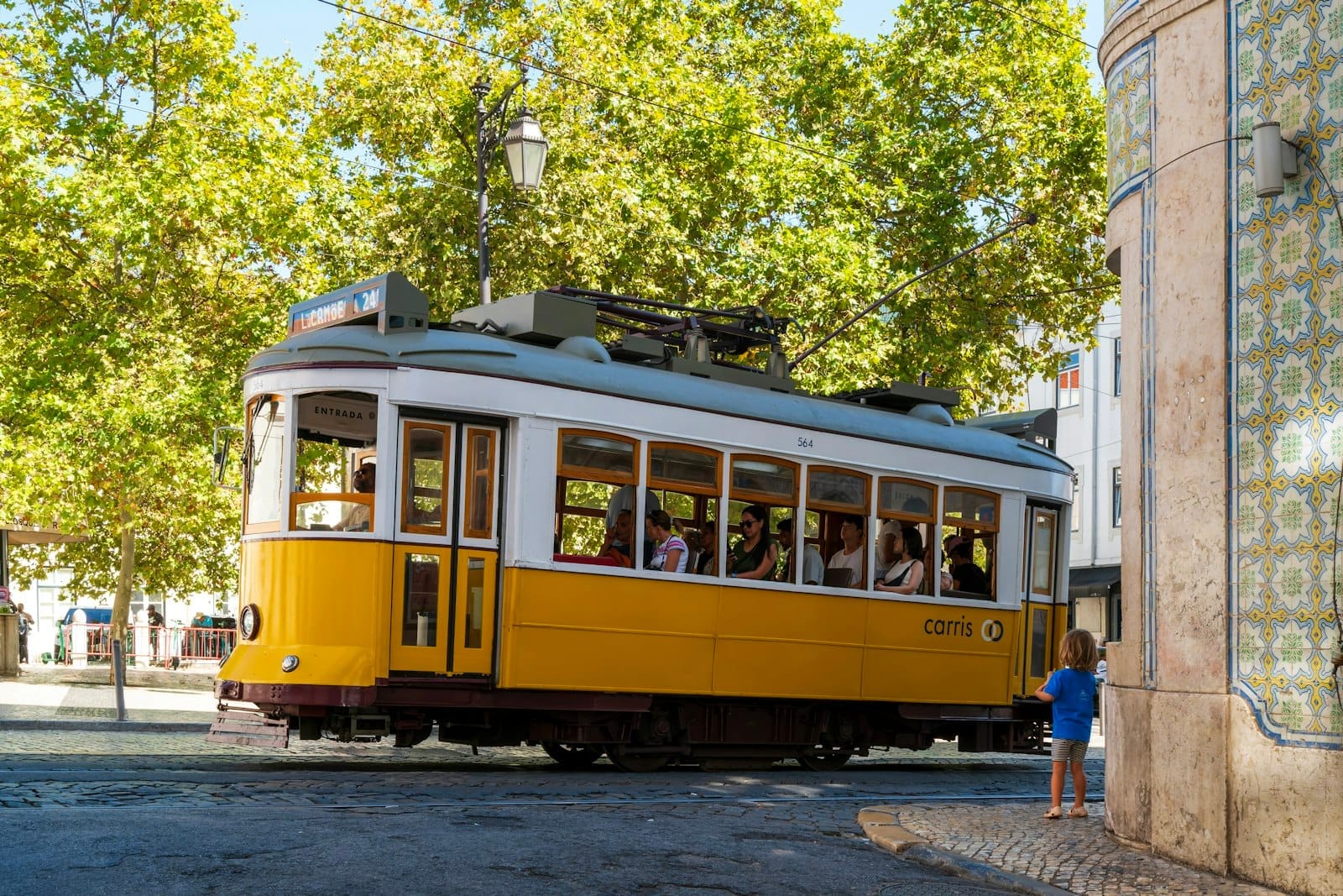Tramway jaune classique tournant à un coin de rue de Lisbonne, avec façade d'immeuble carrelée et arbres feuillus en arrière-plan, capturant l'authenticité de la vie urbaine.