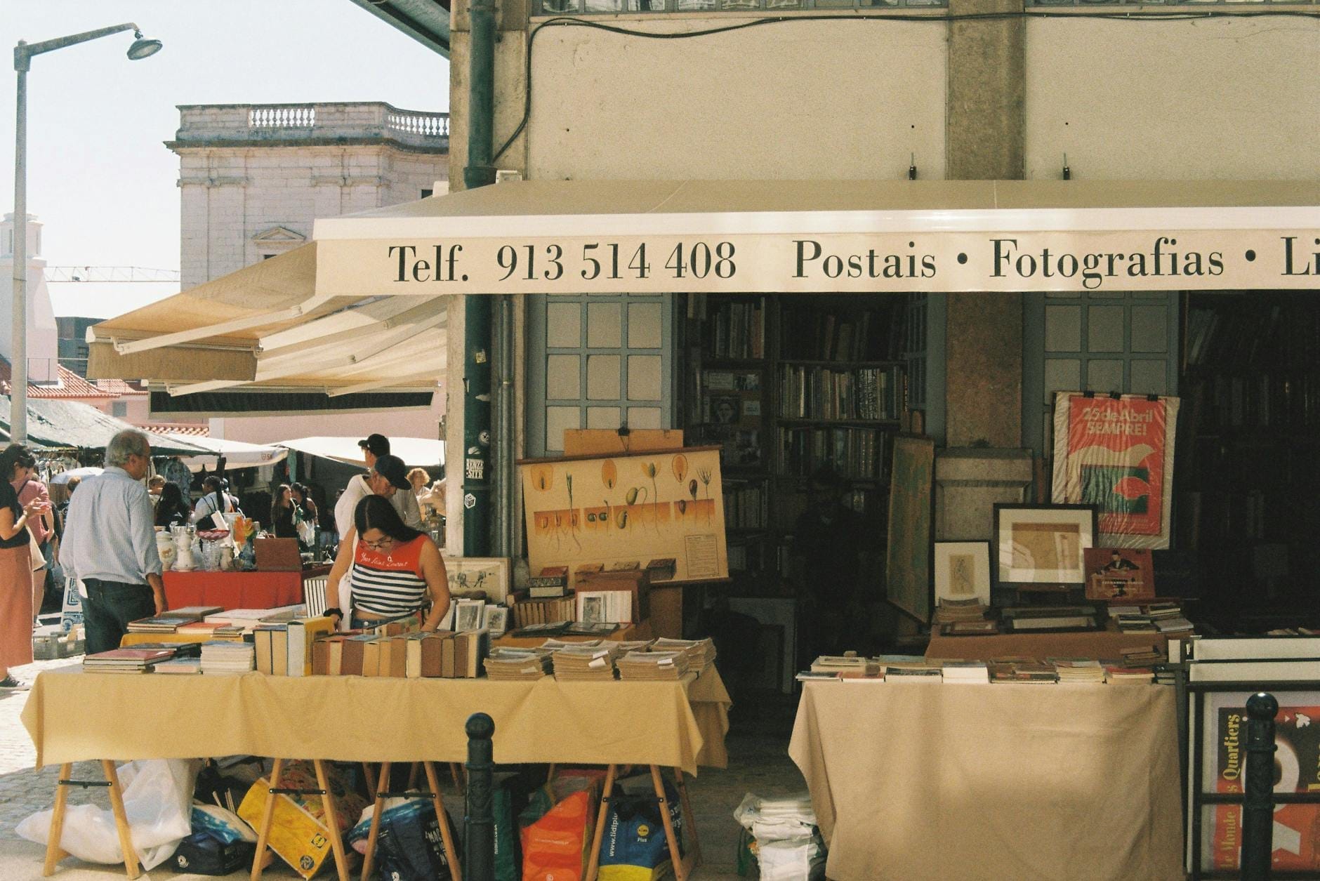 Stand de marché en plein air vendant de vieux livres, cartes postales et photographies à Lisbonne, avec des personnes qui parcourent les articles sous une auvent par une journée ensoleillée.