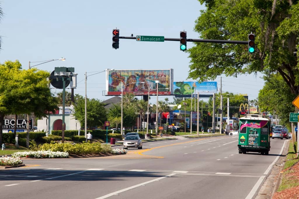 Daytime view of International Drive in Orlando with traffic, green trees, colorful billboards, and visible hotel and restaurant signs under clear skies.