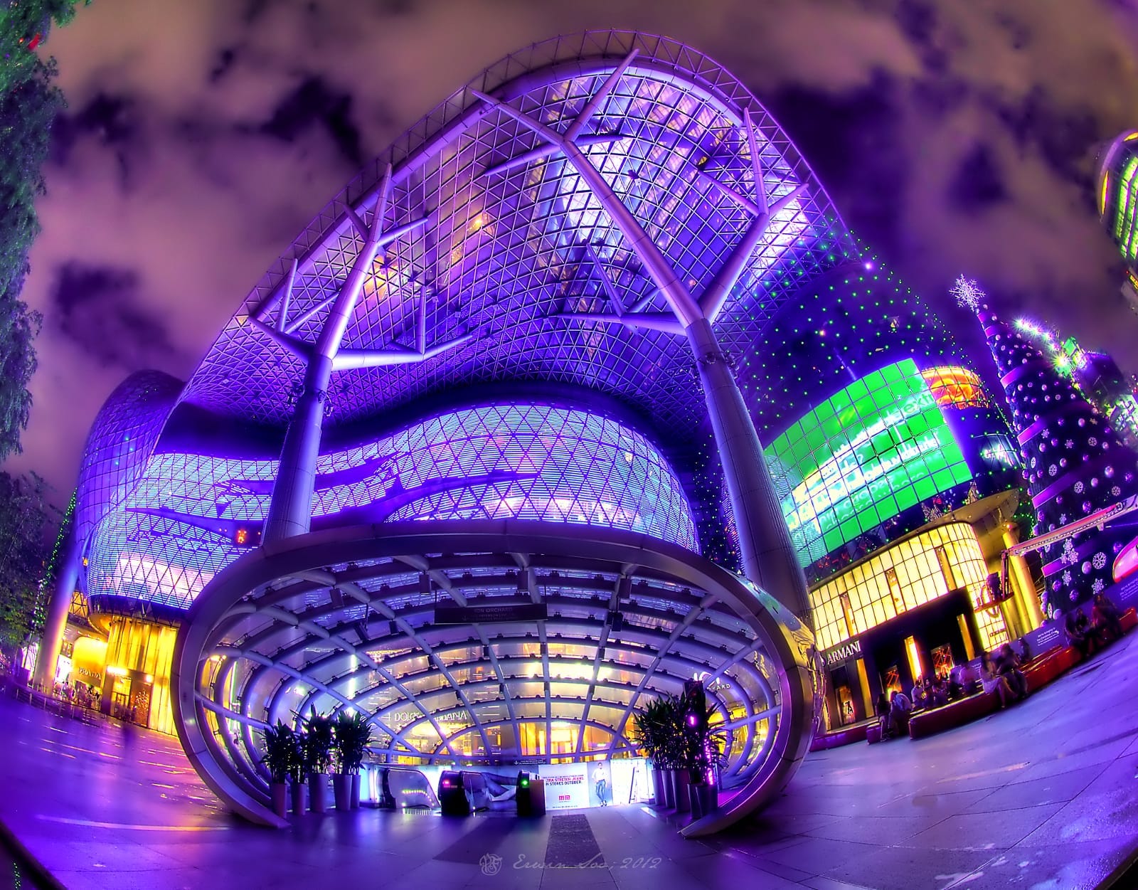 มุมมองแบบไวด์แองเกิลของด้านนอกอาคาร ION Orchard ที่มีดีไซน์ล้ำสมัยด้วยกระจกและเหล็กกล้า ส่องสว่างด้วยแสงสีม่วง น้ำเงิน และเขียวในยามค่ำคืน ดึงดูดนักท่องเที่ยวบน Orchard Road