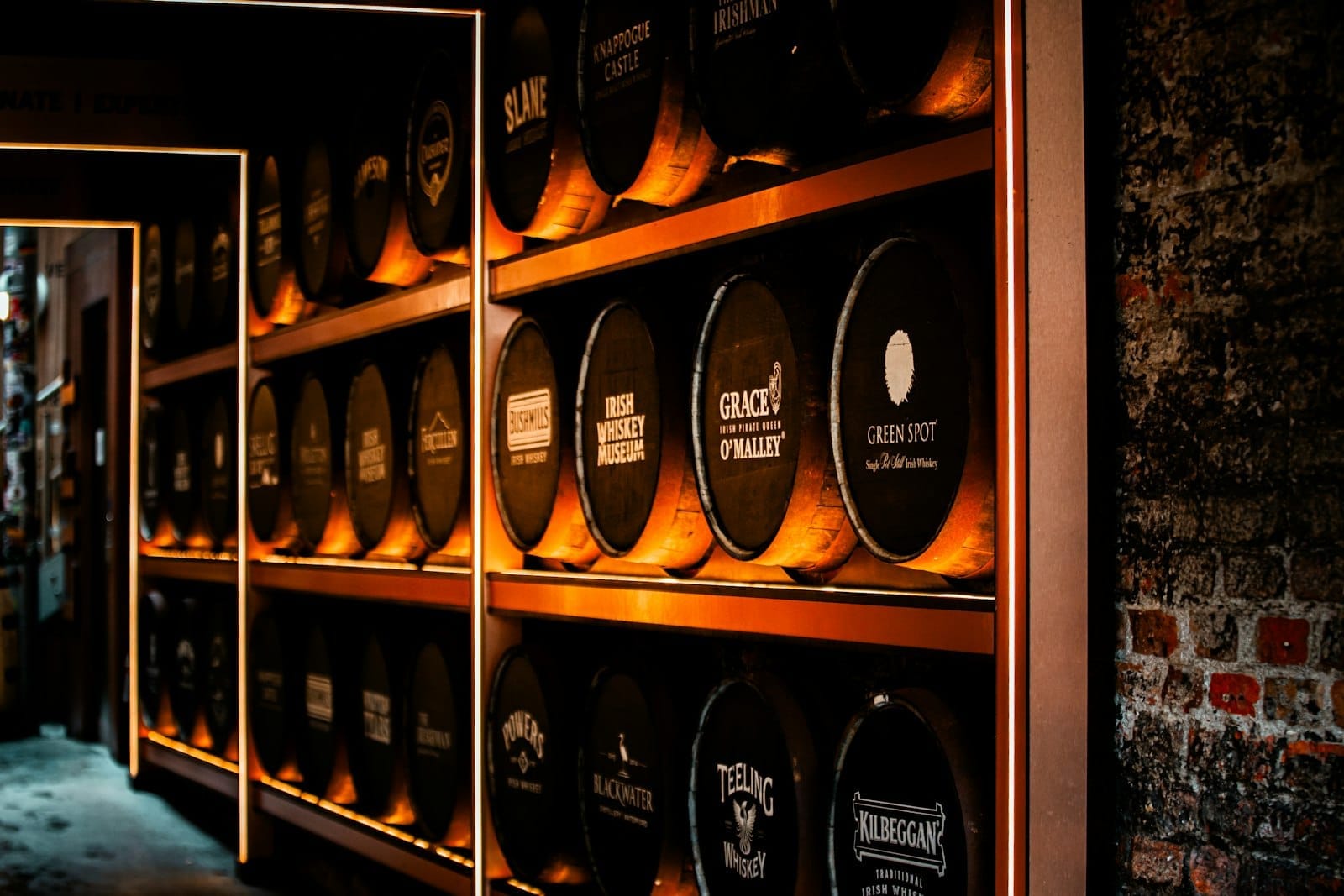 Una parete di botti da whiskey illuminate da una luce calda, con i nomi di vari marchi di whiskey irlandese, all'interno di un museo o distilleria a Dublino.