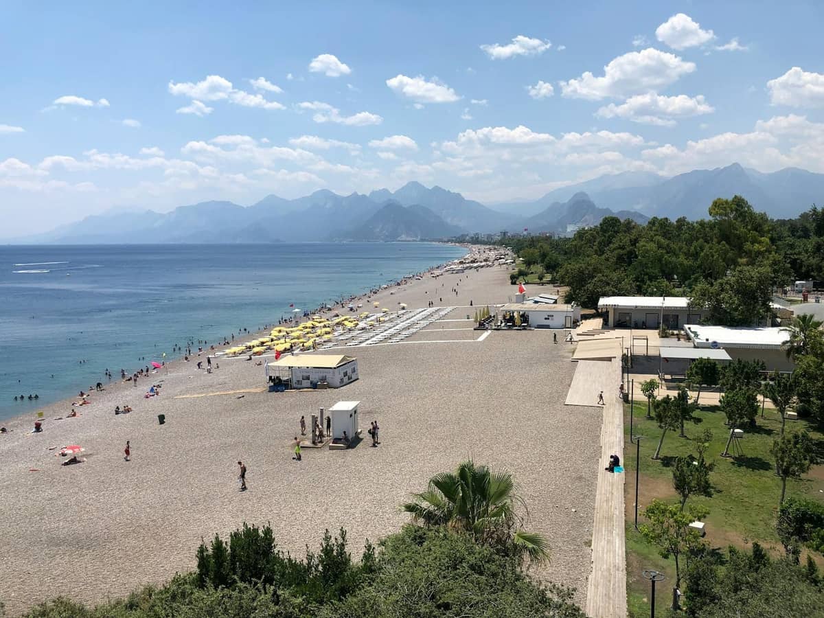 A pebbly Antalya beach with people, sun loungers, nearby park greenery, and dramatic Taurus Mountains in the background.