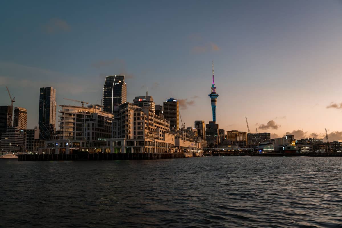 Das Hafenviertel von Auckland in der Dämmerung mit Skyline und Sky Tower, modernen Gebäuden entlang des Hafens und Wasser im Vordergrund.
