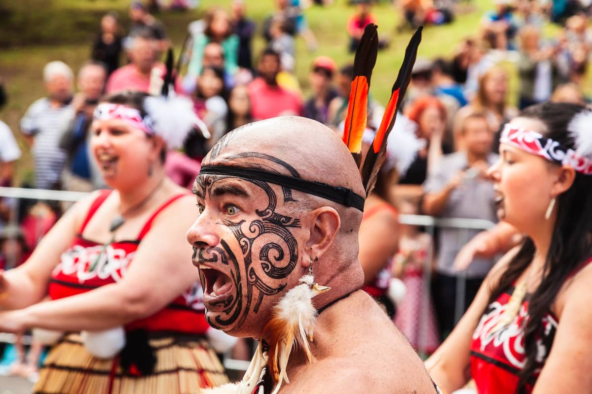 Gruppe māorischer Darsteller in traditioneller Kleidung mit Gesichts-Moko, die an einer Kulturveranstaltung mit einer Menschenmenge im Hintergrund teilnehmen.