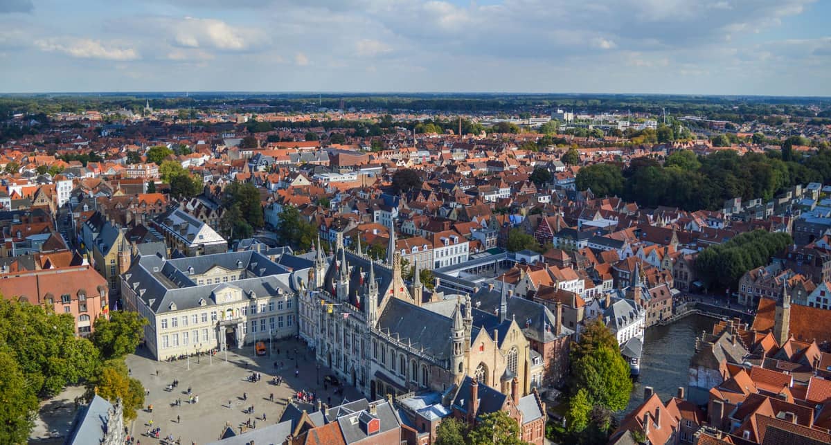 Aerial view of Bruges’ medieval city center showing the main square, historic buildings, red rooftops, and picturesque canals under a blue sky.