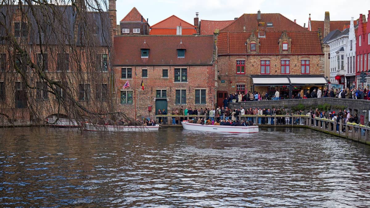 Crowds of people standing in line next to canal boats at Rozenhoedkaai in Bruges with historic brick buildings in the background.