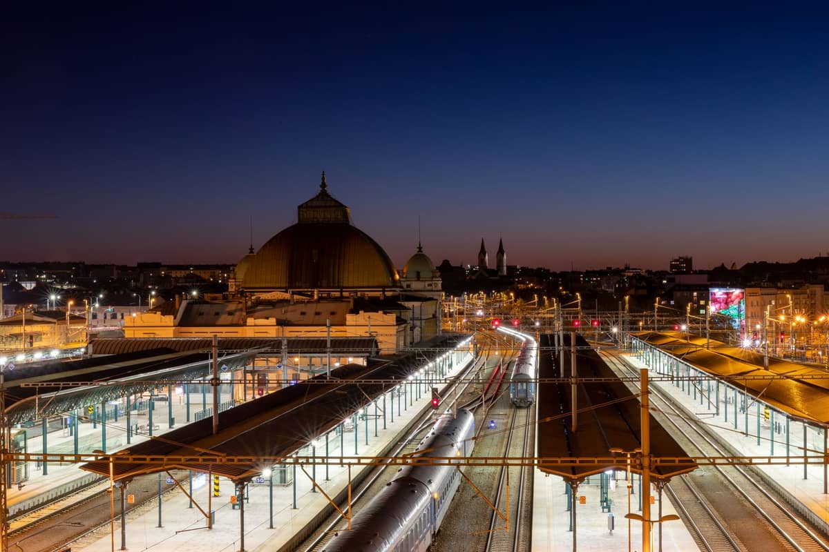 View of a large European train station at dusk with domed roof, platforms, illuminated tracks, and city skyline in the background.