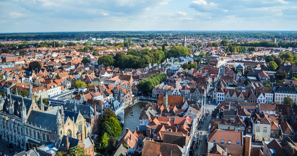 Wide aerial view of Bruges showing the historic center with red-tiled roofs, canals, and medieval buildings under a blue sky.