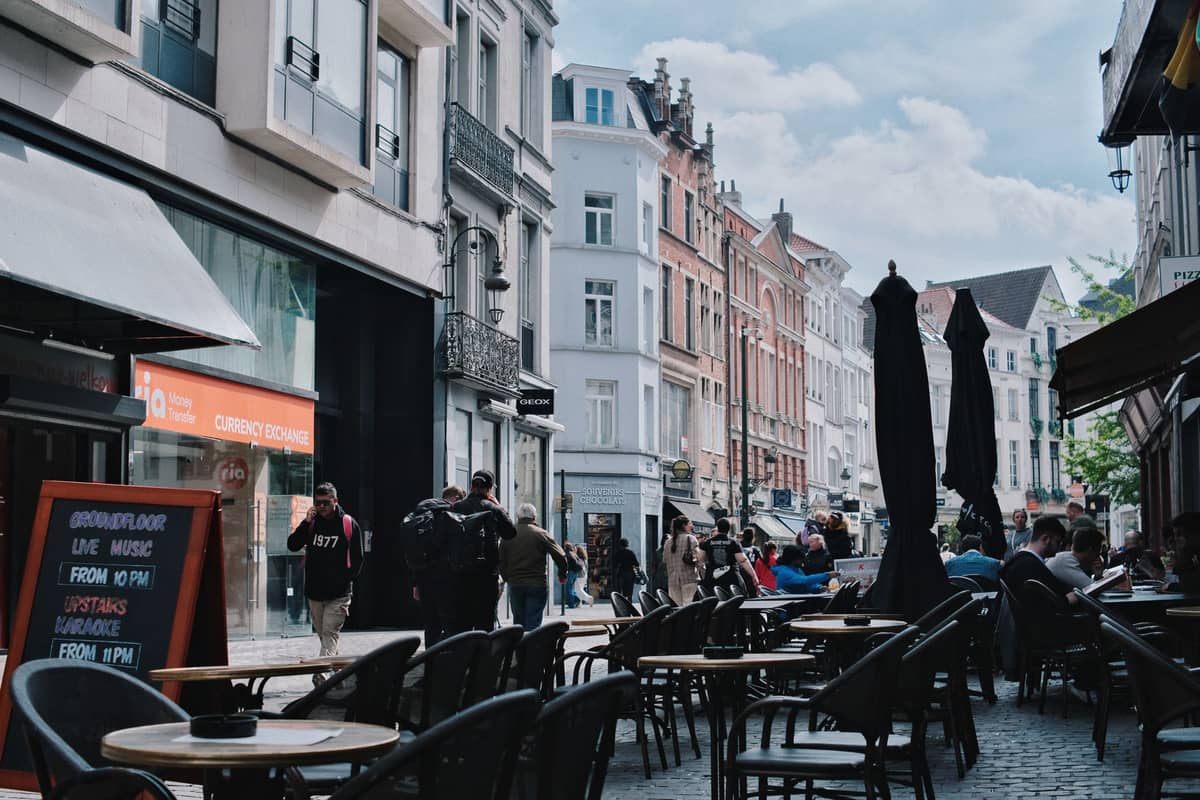 Esplanada de café numa rua de paralelepípedos em Bruxelas com mesas ao ar livre, pessoas a caminhar e arquitetura belga clássica sob um céu levemente nublado