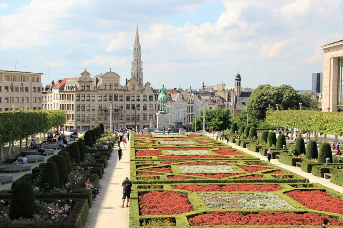 Vista dos jardins do Mont des Arts em Bruxelas, com sebes aparadas, canteiros de flores, pessoas relaxando e edifícios históricos sob um céu ensolarado.