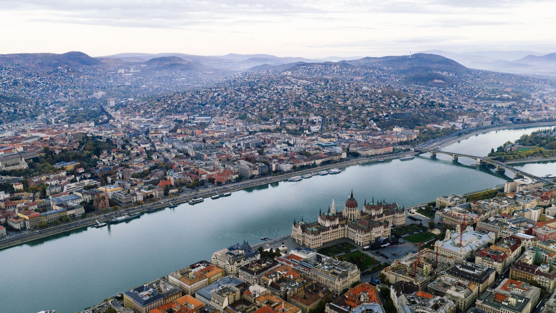 Vista aerea di Budapest con il palazzo del Parlamento, il fiume Danubio, i ponti e un'ampia distesa urbana sullo sfondo di colline lontane.