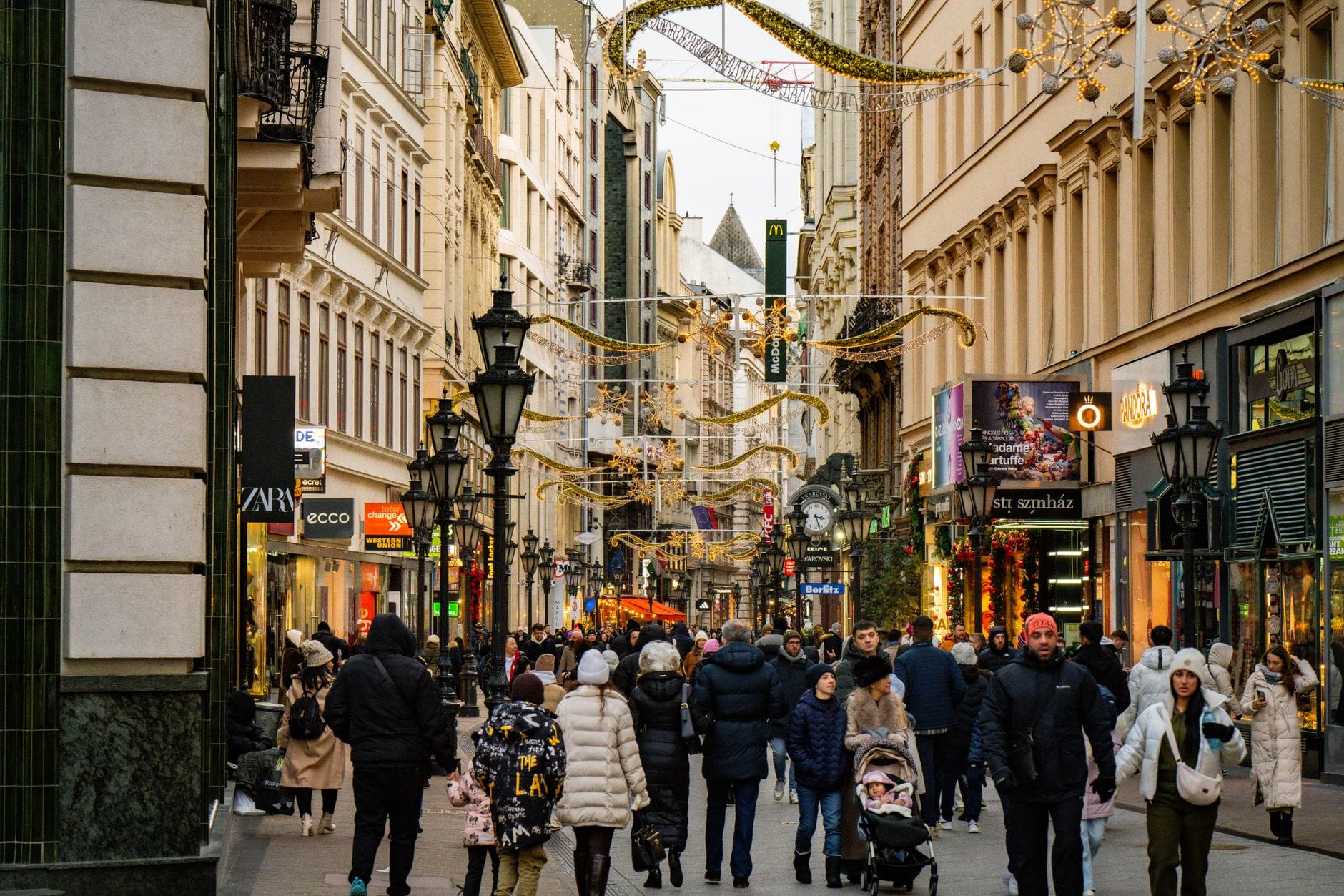 Affollata strada pedonale dello shopping a Budapest fiancheggiata da negozi di souvenir e negozi di marca, con cappotti invernali e luci festive in alto.
