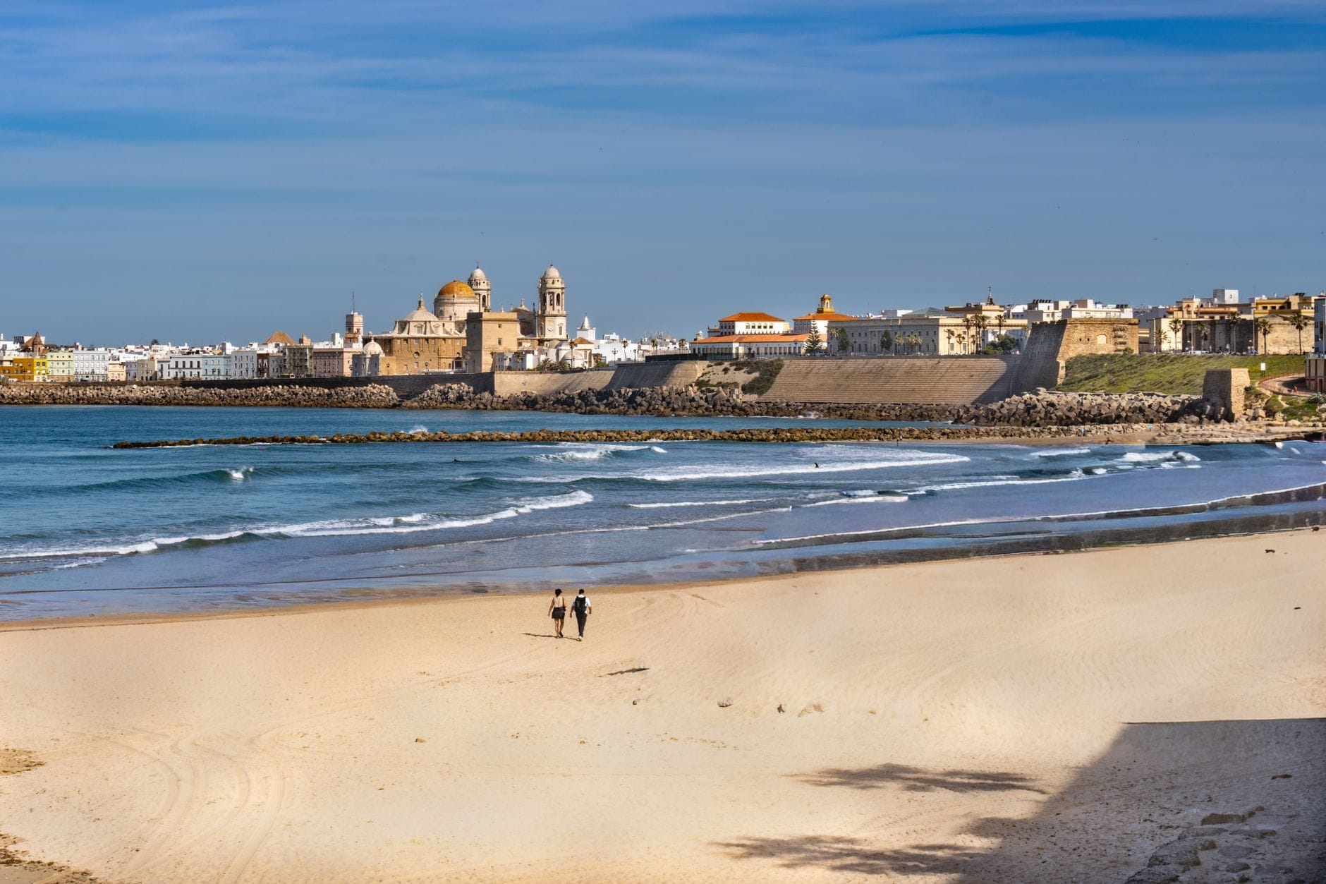 Una spiaggia di sabbia chiara con due persone che camminano, le onde che si infrangono sulla riva, e il centro storico di Cádiz con la cattedrale e le mura sul lungomare sullo sfondo, in una giornata di sole.