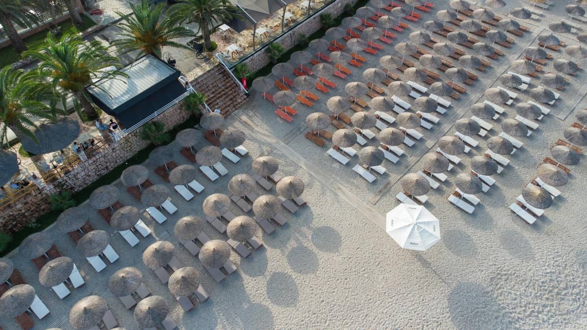 Vista aerea di file di lettini e ombrelloni organizzati su una spiaggia sabbiosa con palme nelle vicinanze, che illustra i club balneari privati.