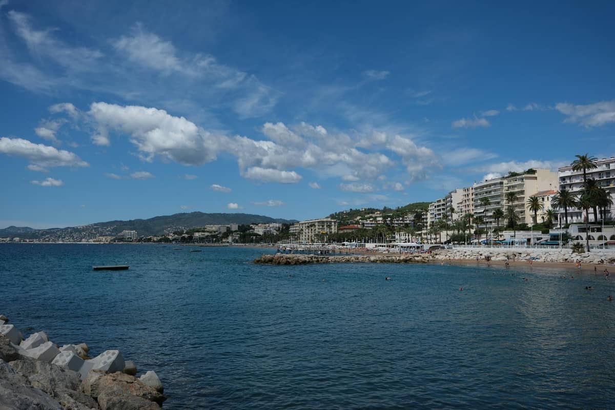 Vista del lungomare di Cannes con acque blu limpide, spiaggia sabbiosa, palme ed eleganti edifici sotto un cielo azzurro brillante.
