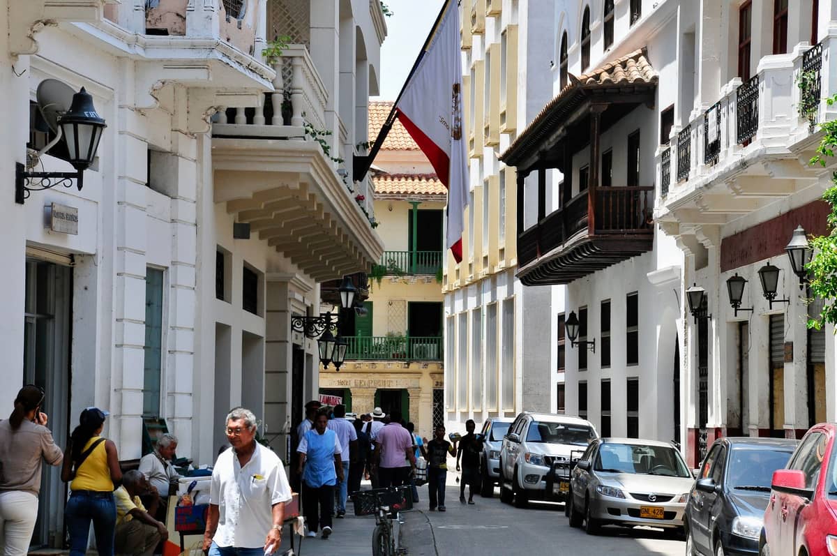 A lively Cartagena street with colonial architecture, people walking and cars parked, capturing daily life in the bustling historic district.