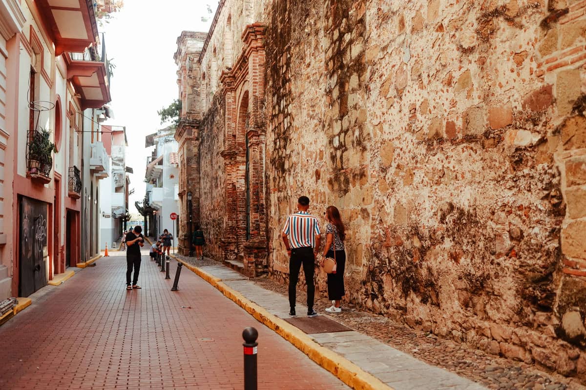 People stand and walk along a narrow street beside an old stone wall in Cartagena’s historic Walled City under bright sunlight.