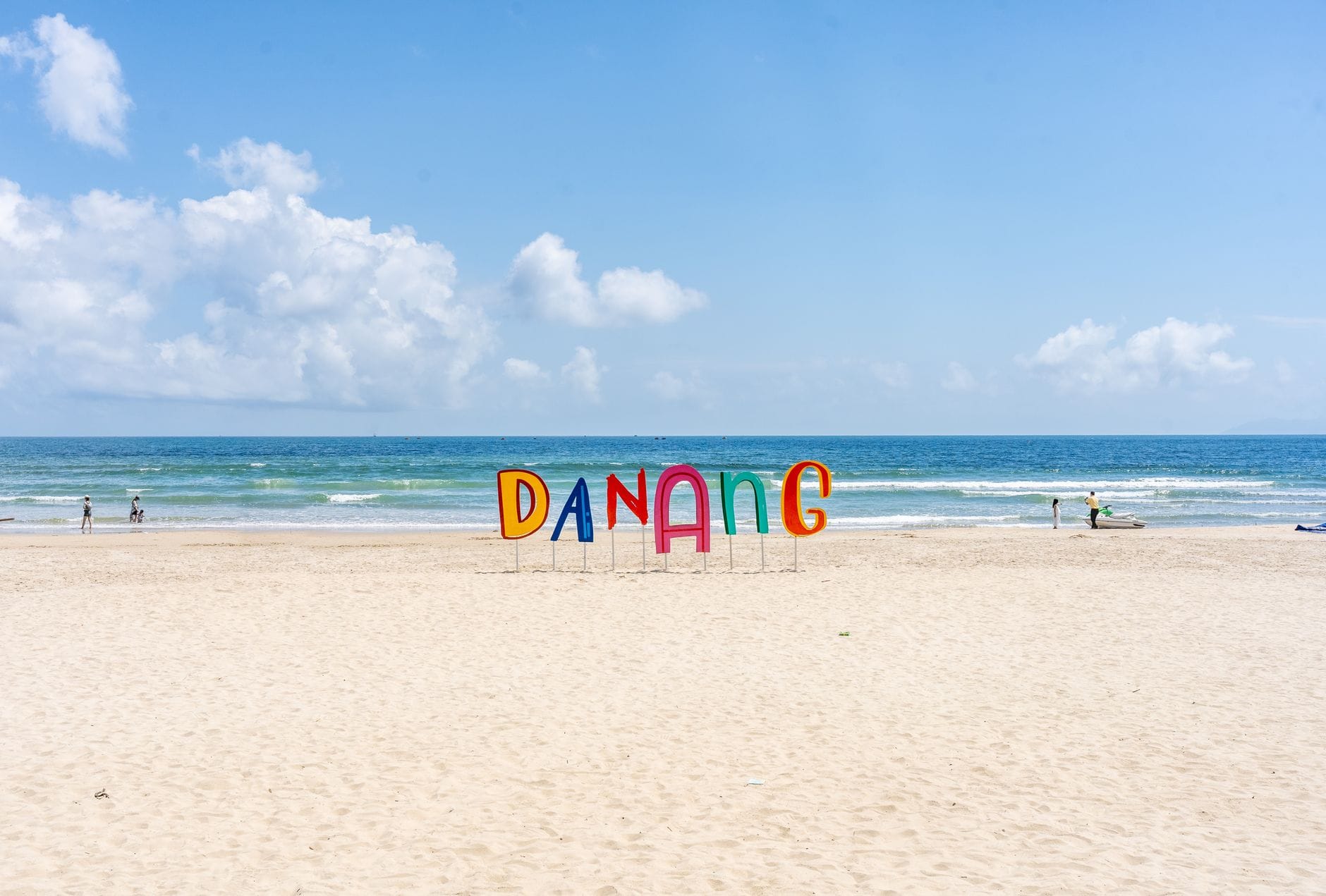 ชายหาดทรายกว้างในดานังพร้อมป้าย 'DA NANG' สีสันสดใส คลื่นเบาๆ ท้องฟ้าสีฟ้า และผู้คนไม่กี่คนในระยะไกลที่เพลิดเพลินกับชายฝั่ง