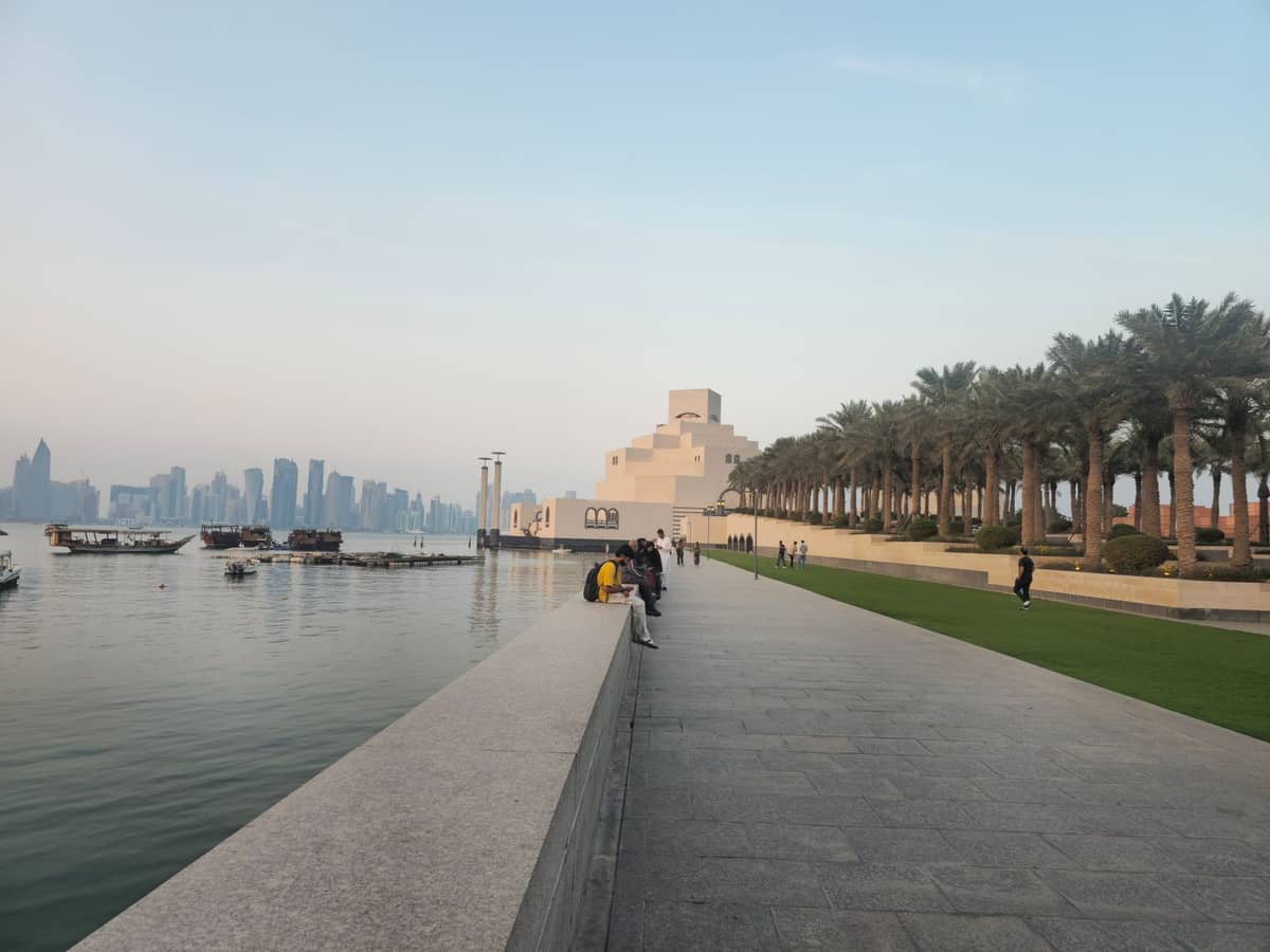 Palmengesäumte Promenade der Doha Corniche mit Spaziergängern, dem Museum für Islamische Kunst und der Skyline der Stadt am Wasser an einem klaren Tag.
