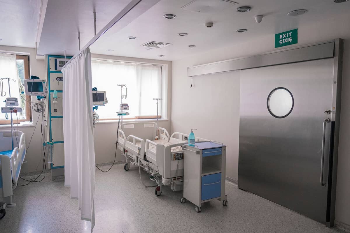 ห้องพักในโรงพยาบาลที่สว่างและทันสมัย พร้อมเตียงทางการแพทย์ อุปกรณ์ตรวจสอบ และประตูโลหะขนาดใหญ่ สื่อถึงสิ่งอำนวยความสะดวกด้านการดูแลสุขภาพที่มีมาตรฐานสูง