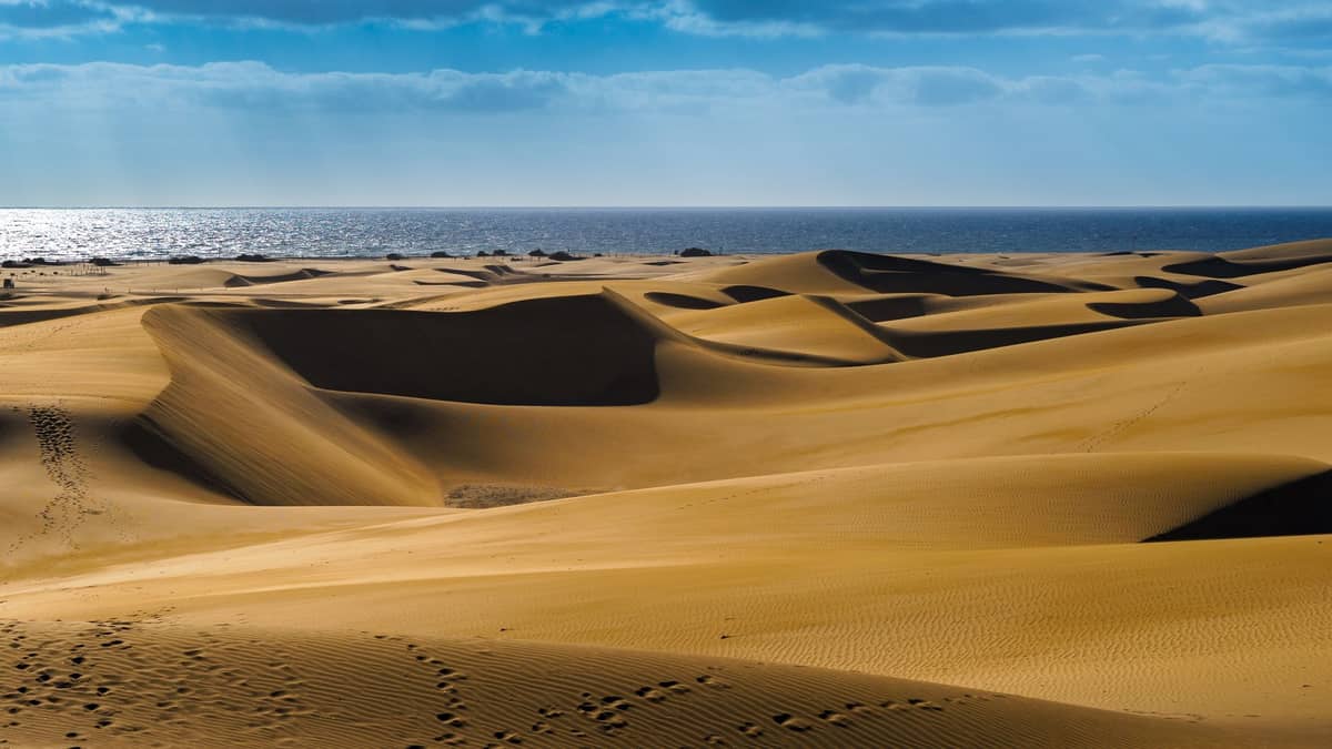 Панорамный вид на золотые песчаные дюны с голубым Атлантическим океаном и частично облачным небом на заднем плане, характерный для Гран-Канарии.